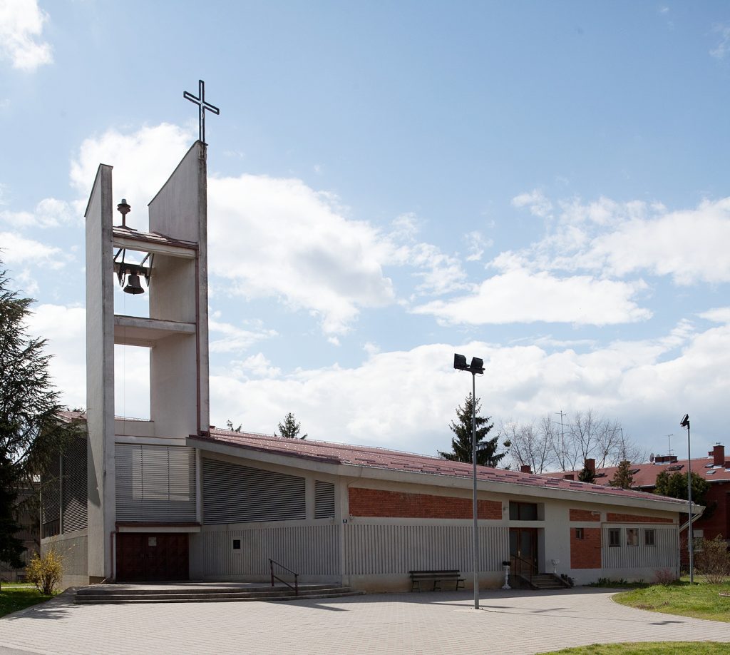 White church building with a tall bell tower and a cross atop, against a blue sky, in daylight.