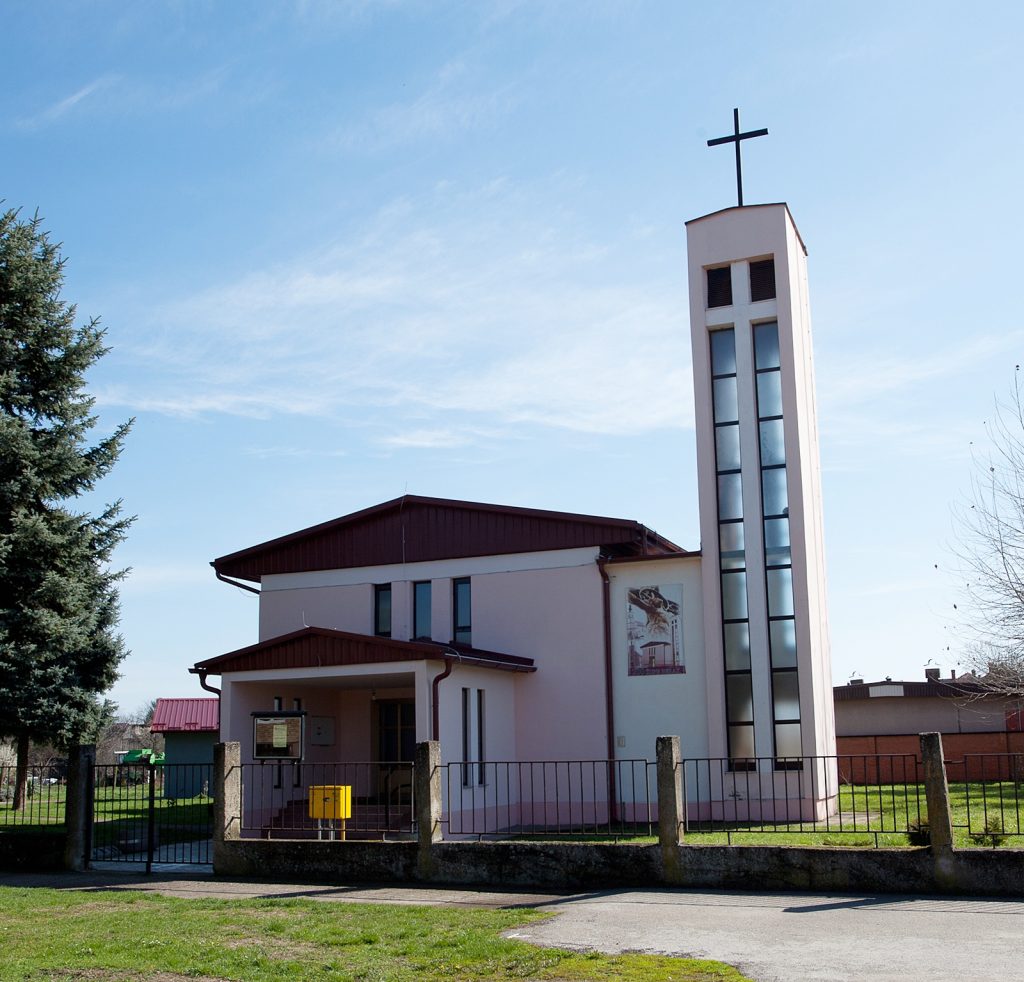 Church building with a tall rectangular bell tower topped by a cross, set against a blue sky and surrounded by a fence and green lawn.