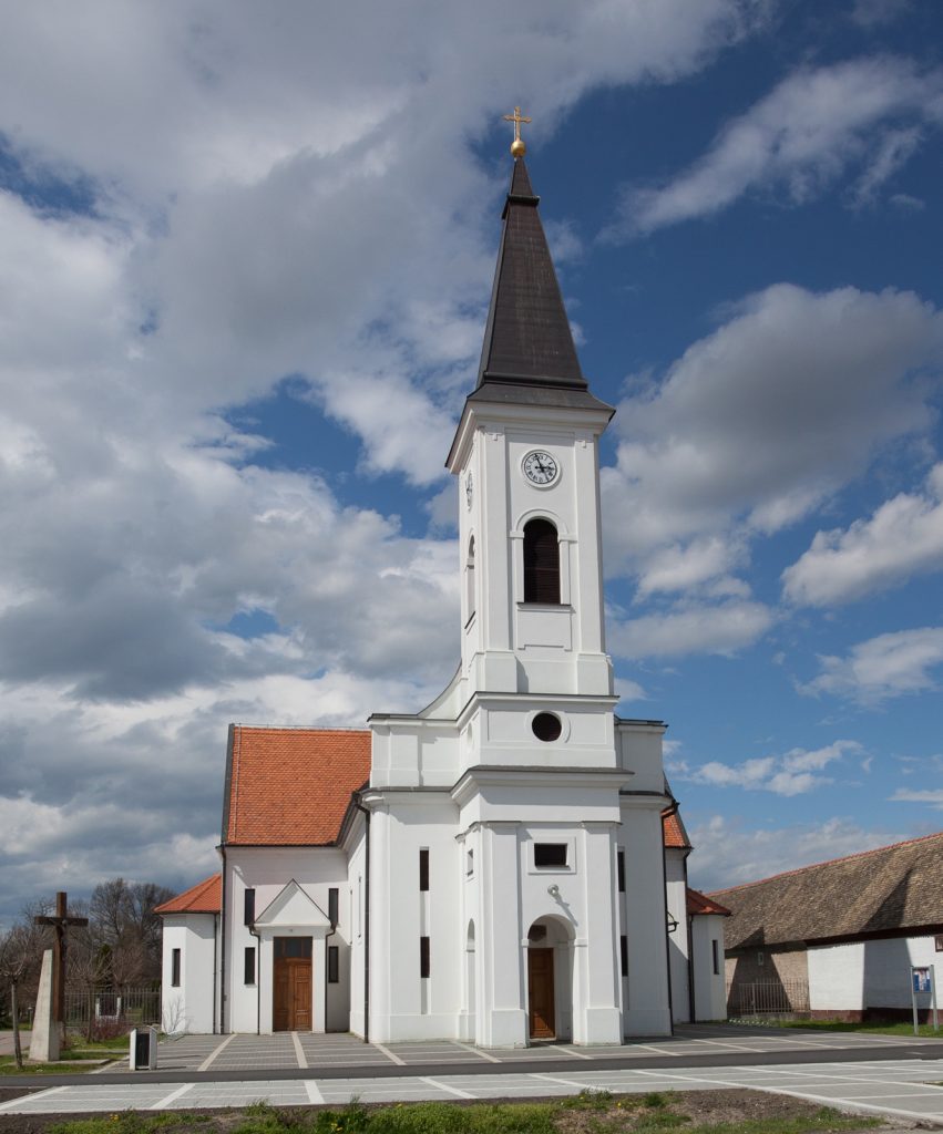 White church with a tall steeple topped by a gold cross against a blue sky with clouds; clock tower visible.