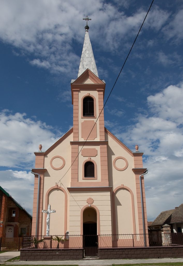 Pink church with a tall silver steeple and cross atop, set against a blue sky with clouds.