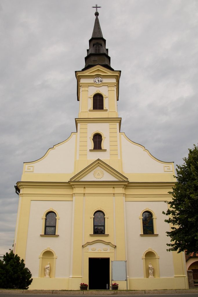 Yellow and white church facade with a tall, multi-tiered steeple and a clock, set against a cloudy sky
