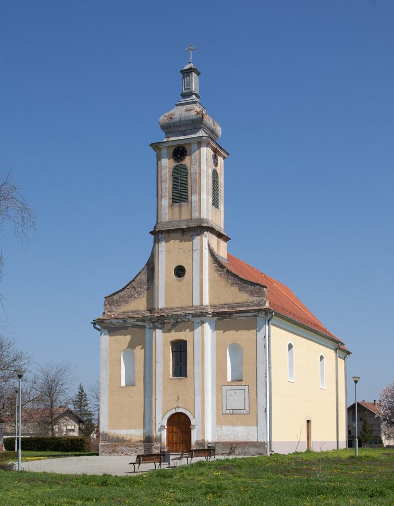 Small yellow church with tall steeple topped by a cross against a clear blue sky, benches outside.