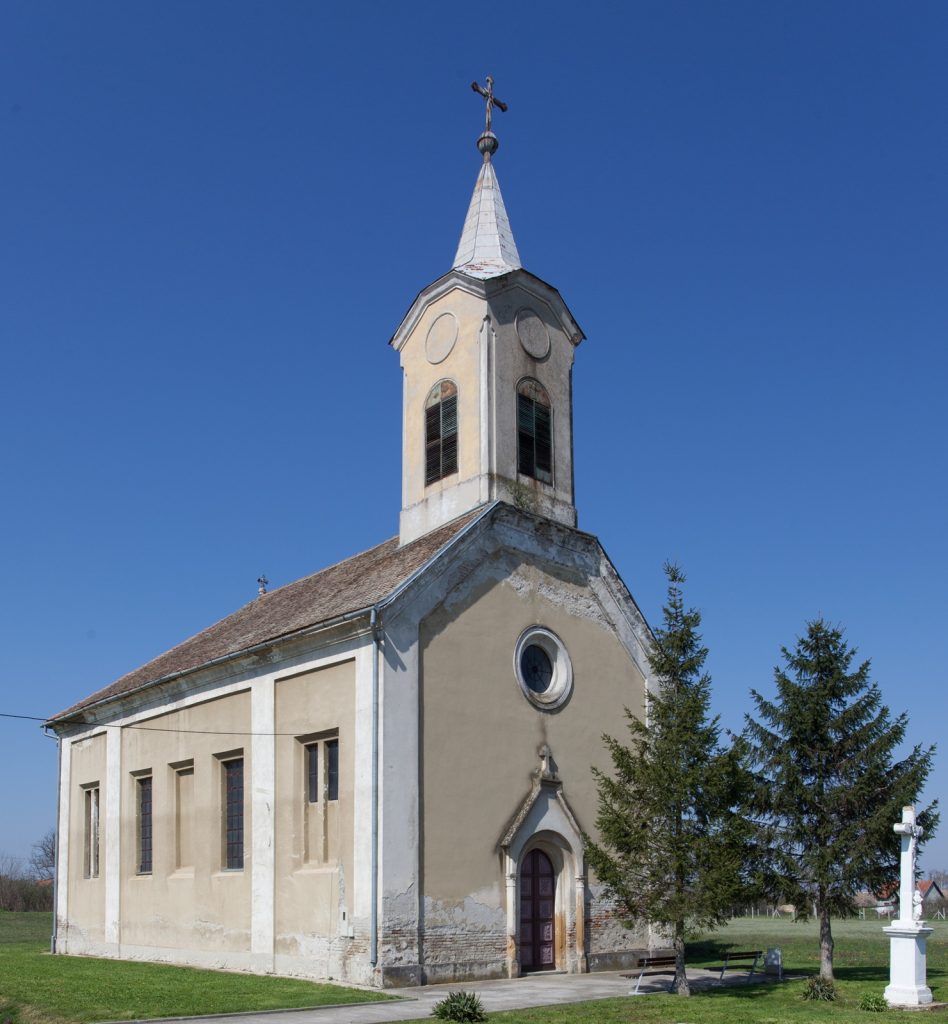 Small church with a steeple and cross against a clear blue sky; beige facade, arched windows, and stone door facade.