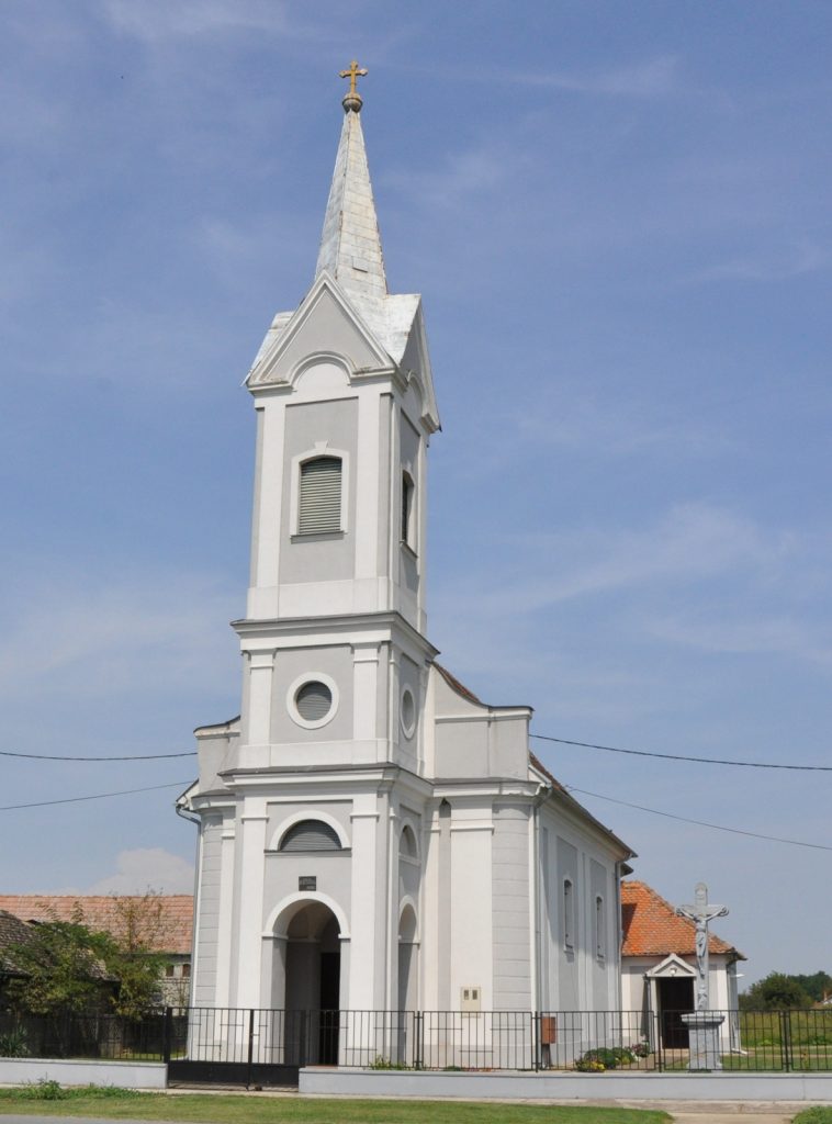 White church with a tall steeple topped by a gold cross against a blue sky, front door archway visible at ground level with a black fence surrounding the building.