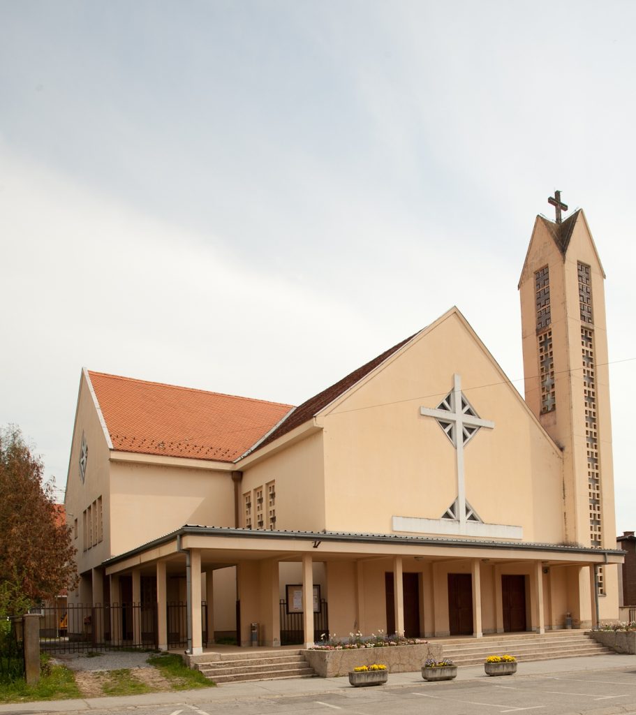 Beige church with red-tiled roof and tall bell tower topped with a cross standing beside a covered entrance.