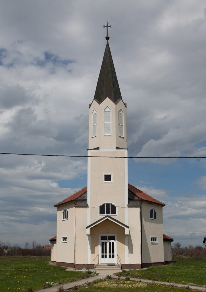 Church building with a tall steeple and cross on top, set in a rural landscape under a cloudy sky