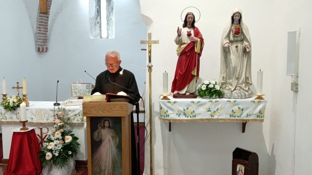 Priest at a wooden lectern reading aloud in a church with an ornate altar behind him and statues of Jesus and Mary nearby.