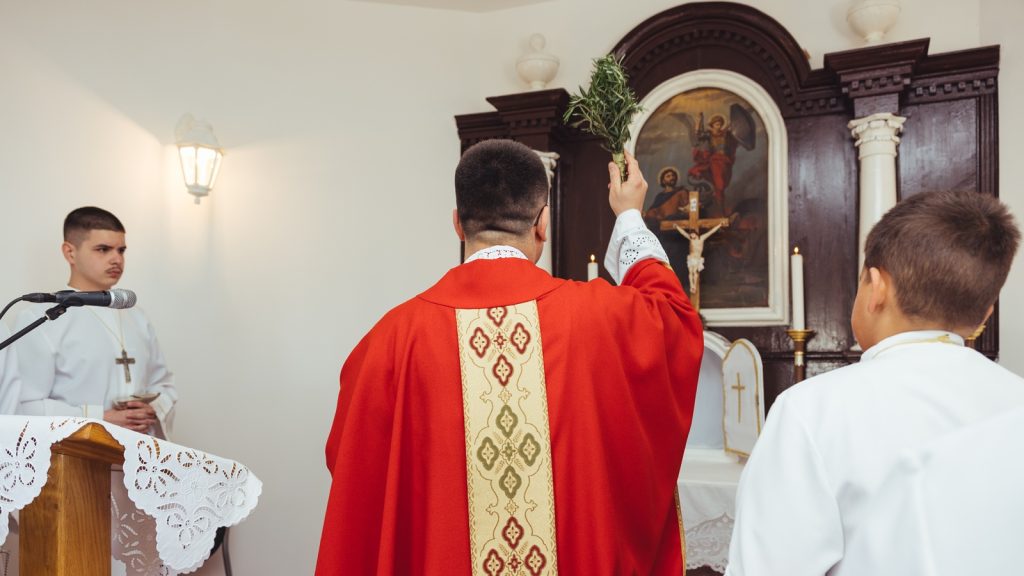 Priest in red vestments sprinkling holy water from a sprig, with two altar servers in white robes nearby at the ornate altar.