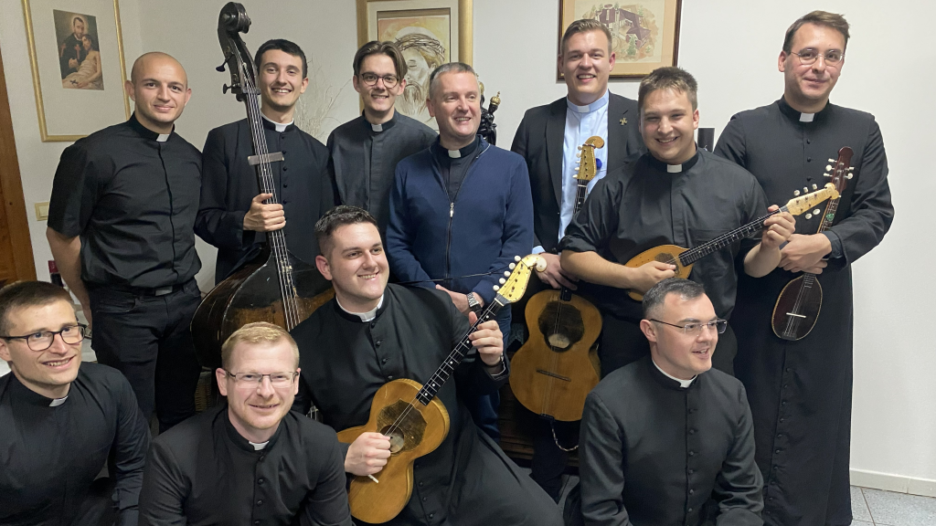 A group of priests in black clerical shirts posing with guitars and a bass in a room with framed art on the walls.