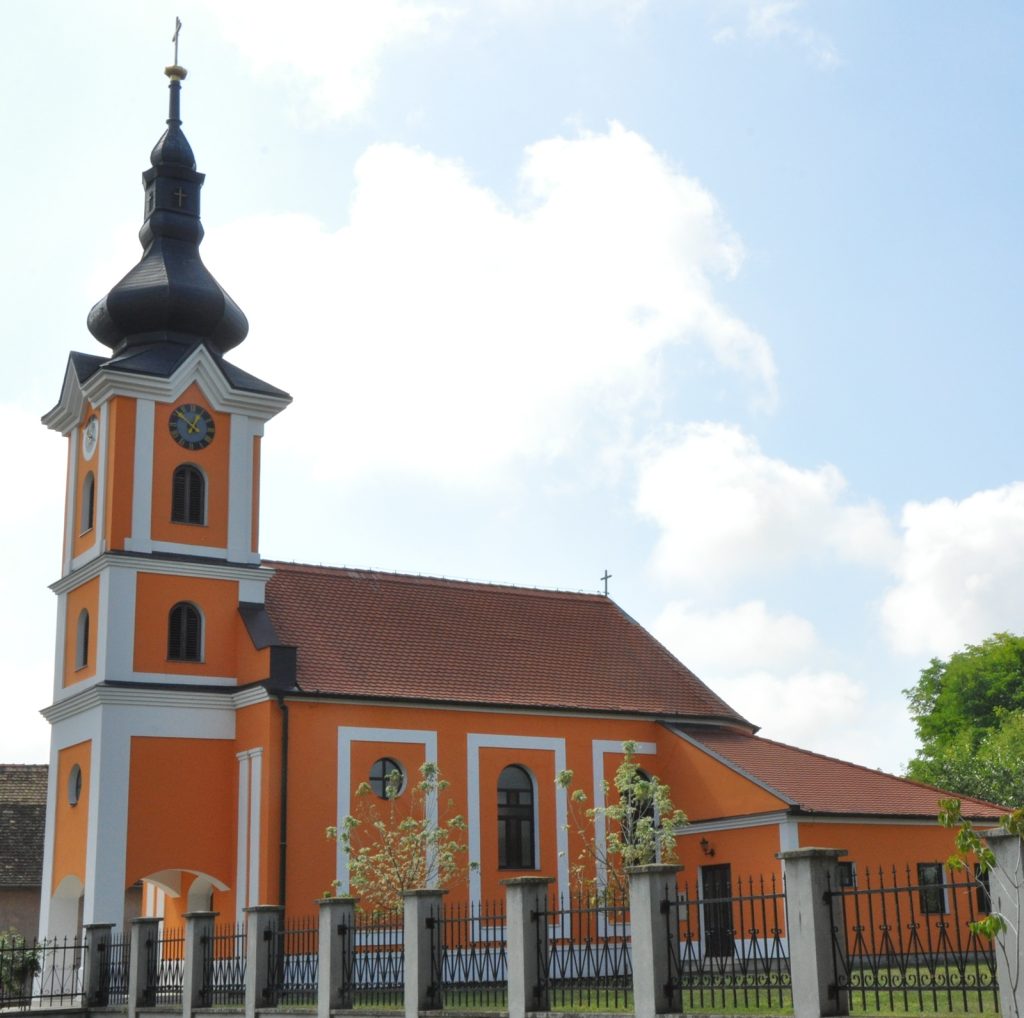 Orange church with white trim and a tall black onion dome steeple, clock faces on the tower, set against a bright blue sky with clouds, bordered by a black wrought-iron fence.