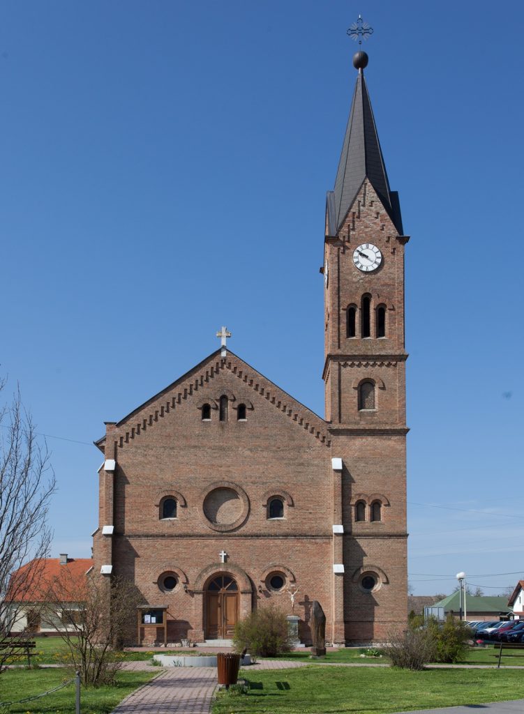 Brick church with a tall clock tower and cross on the roof, set against a clear blue sky.