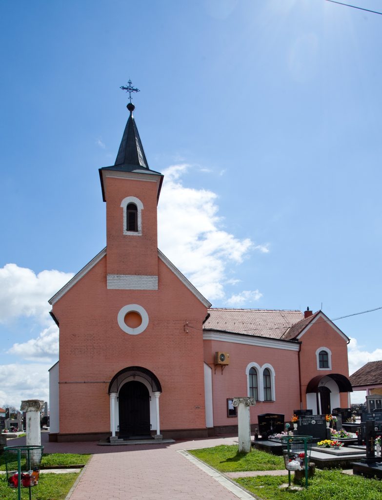 Pink brick church with a tall steeple and cross under a blue sky; cemetery graves visible in the foreground.