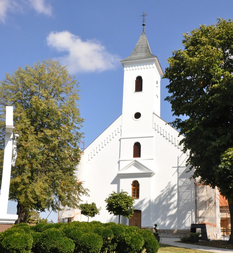 White church with tall steeple against a blue sky, surrounded by green trees and trimmed hedges, front door visible.