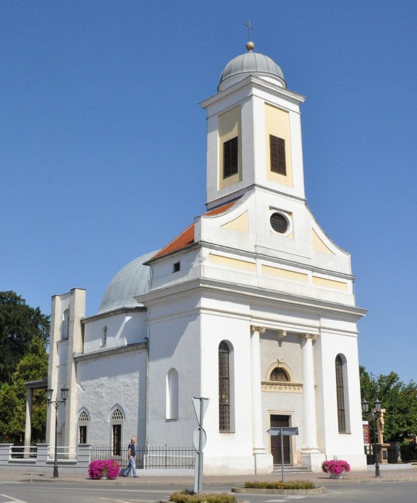 White church with a tall bell tower, dome and cross against a clear blue sky, yellow-trimmed façade nearby.