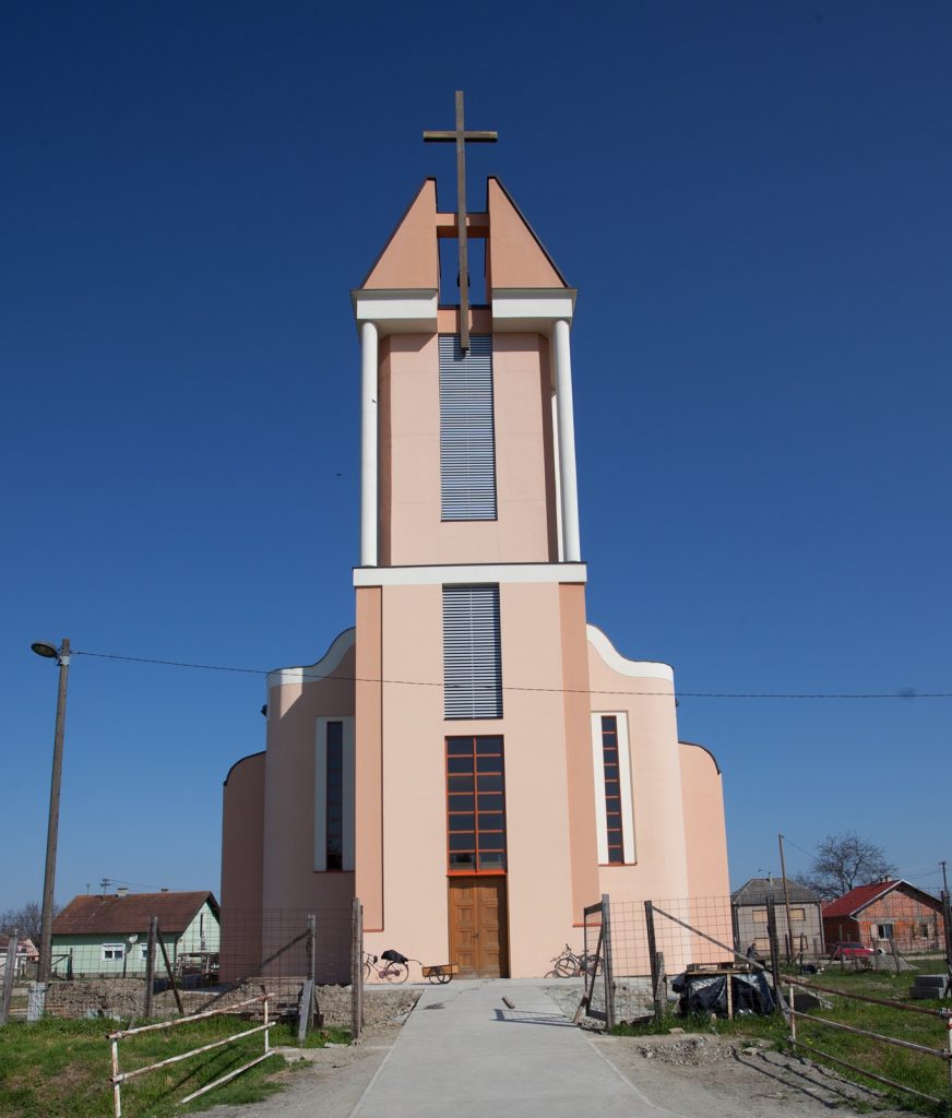 Pink church tower with a tall cross atop, set against a clear blue sky; path leads to the wooden doors at the base.
