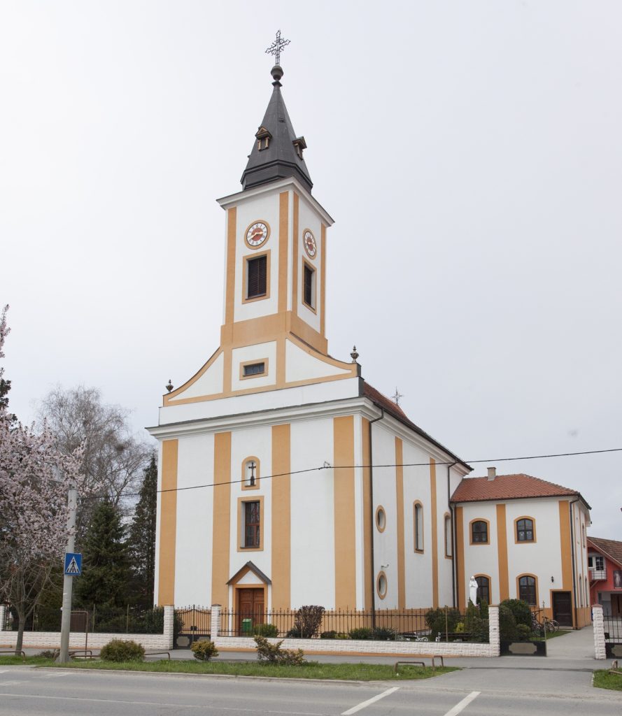 Front view of a pale yellow church with orange trim, a tall clock-tower, and a dark gray spire topped with a cross against a pale sky.