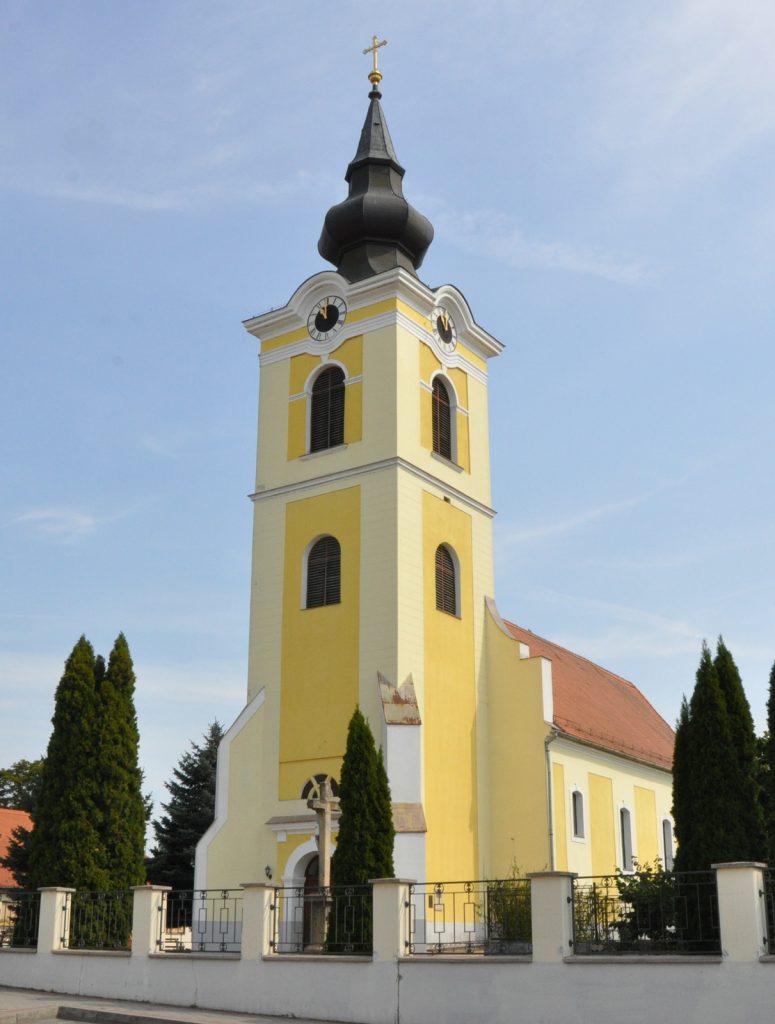 Yellow church tower with two clock faces and a dark onion-shaped spire topped by a gold cross, against a blue sky.