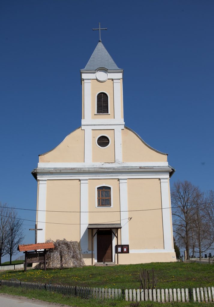 Yellow church with white trim and a tall steeple topped by a cross against a blue sky. | Informative still image of a church exterior with steeple and cross flourish on top.