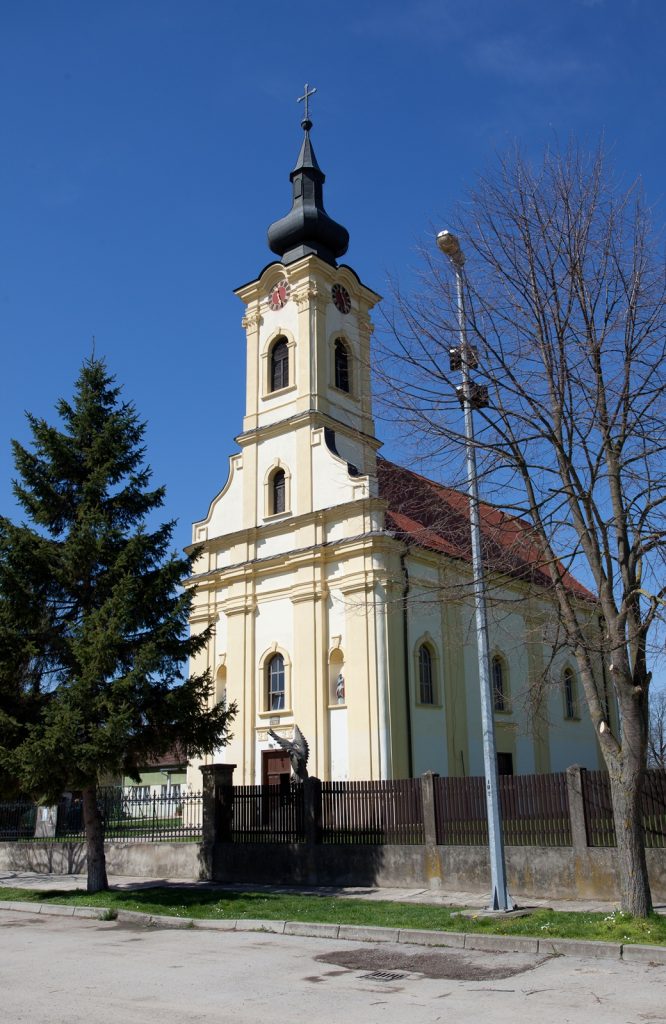Front view of a pale yellow church with a tall clock-tower, dark onion dome, and cross atop, against a clear blue sky.