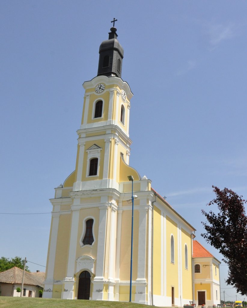 Yellow church with a tall bell tower and cross, clock faces on the tower, against a clear blue sky.