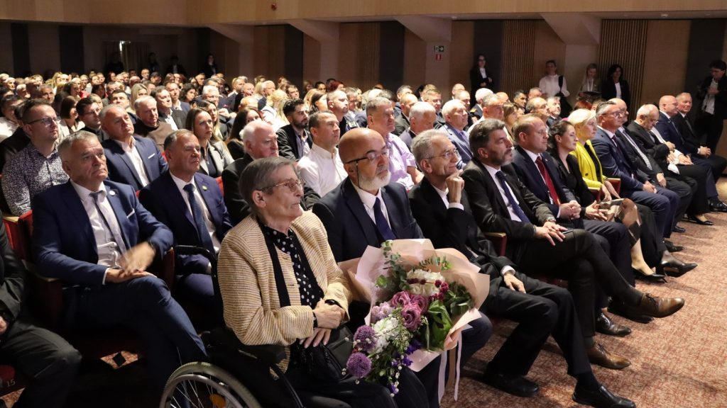 Audience gathered in a formal hall, rows of people in suits watching a ceremony; foreground includes a man in a wheelchair holding a bouquet of flowers.