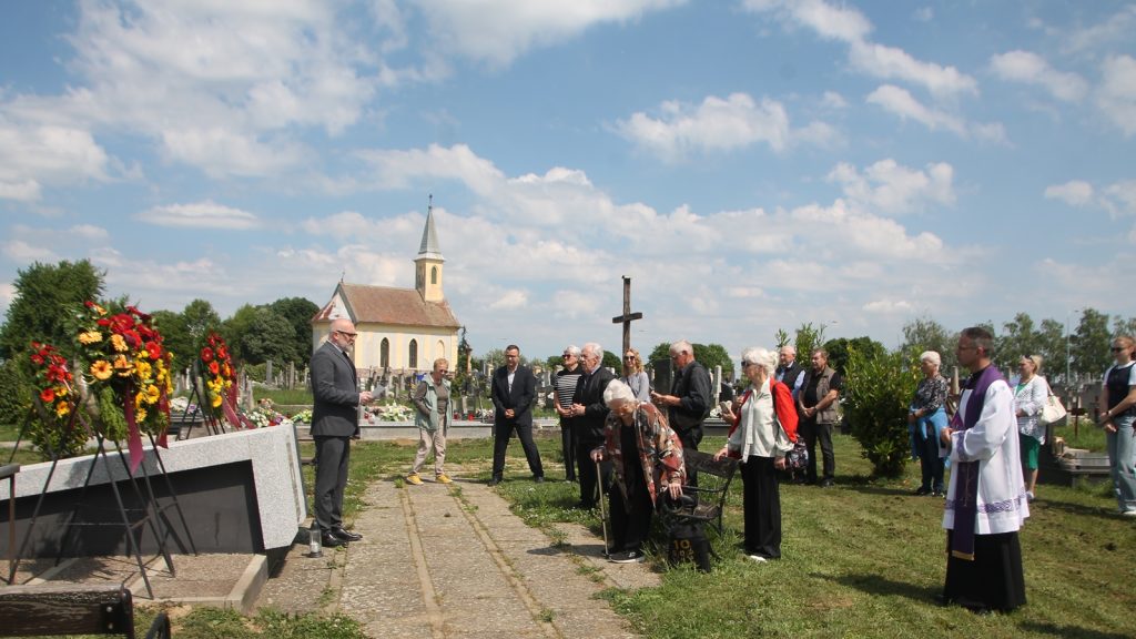 A group of people gathered in a cemetery for a memorial ceremony, with a priest in white robes and a church in the background on a sunny day.