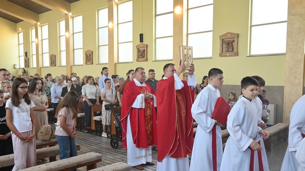 Priest in red raises a gilded book during a church service as altar servers in white robes stand nearby.