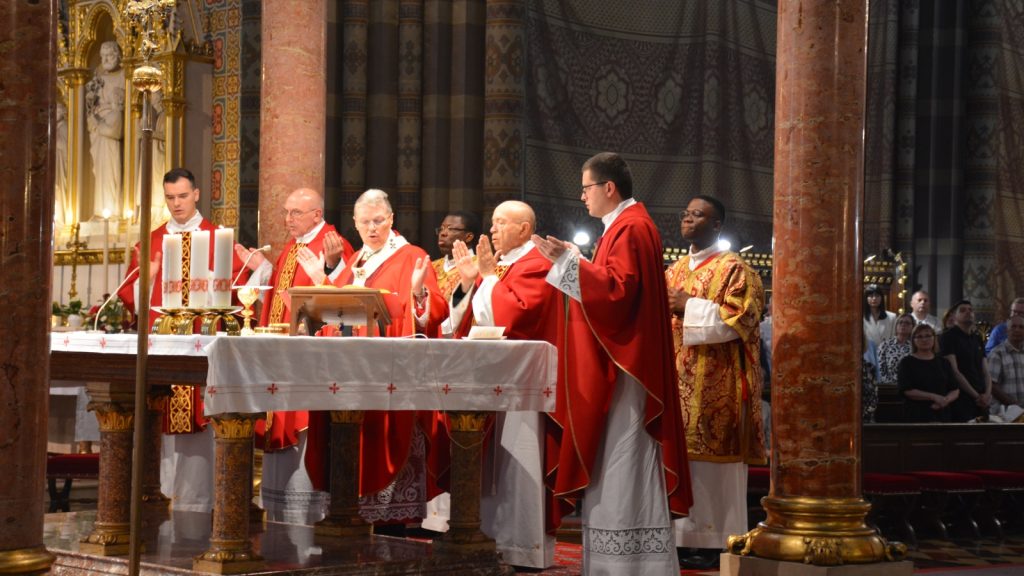 Catholic priests in red vestments celebrate Mass at an ornate altar inside a decorative church.