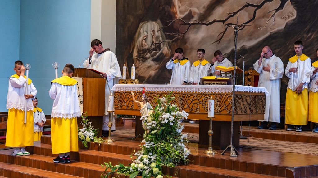 Priest at a wooden lectern delivers a sermon during a Mass, surrounded by altar servers in white and yellow robes at an ornate altar.