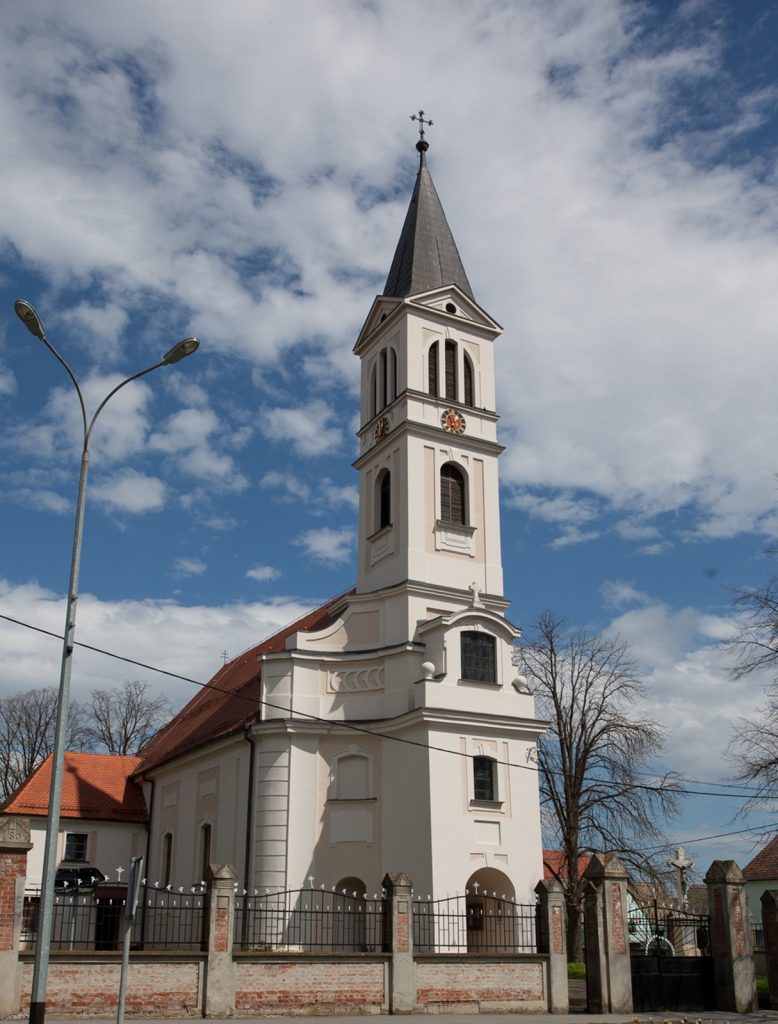 White church with a tall, pointed steeple and clock faces, set against a blue sky with clouds; a curved street lamp in the foreground.
