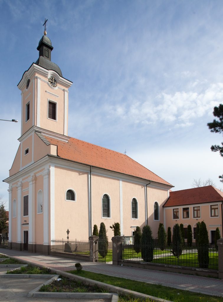 Peach-colored church with a tall clock tower and red-tiled roof under a blue sky, surrounded by a black fence.