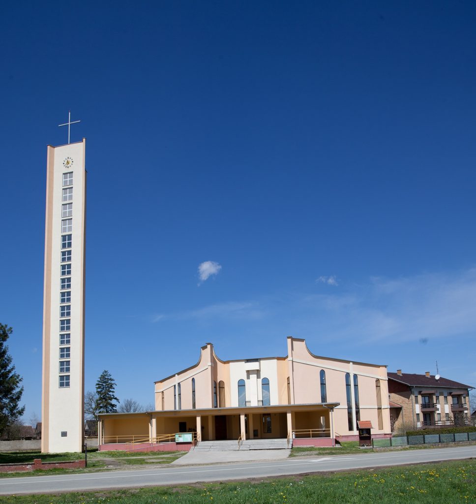 Tall clock tower with a cross on top beside a peach-colored church building under a bright blue sky.