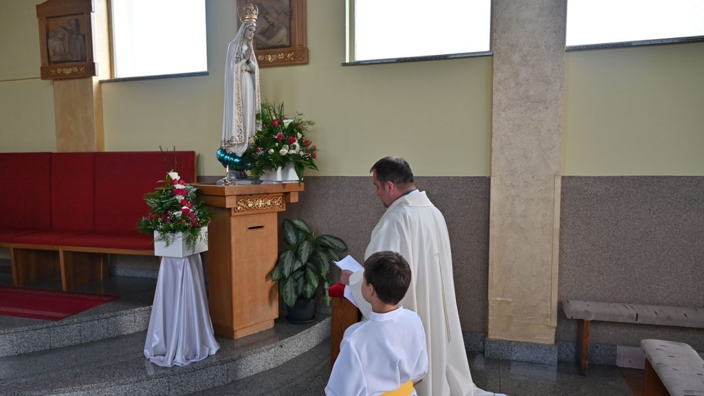 Priest and a young altar server kneel in prayer before a statue of the Virgin Mary with flowers in a church setting.