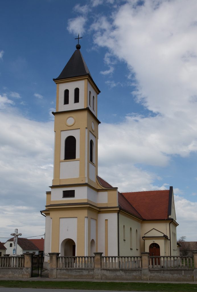 Front view of a pale yellow and white church with a tall steeple and cross against a blue sky with clouds, surrounded by a low stone fence