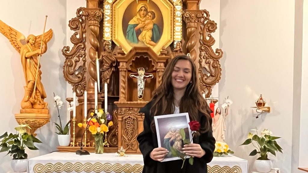 Young woman smiling in a church, holding a printed religious image and a red rose, in front of an ornate altar.