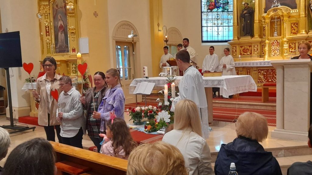 Children stand in a church aisle holding red heart decorations during a service, with altar and clergy in white robes behind them