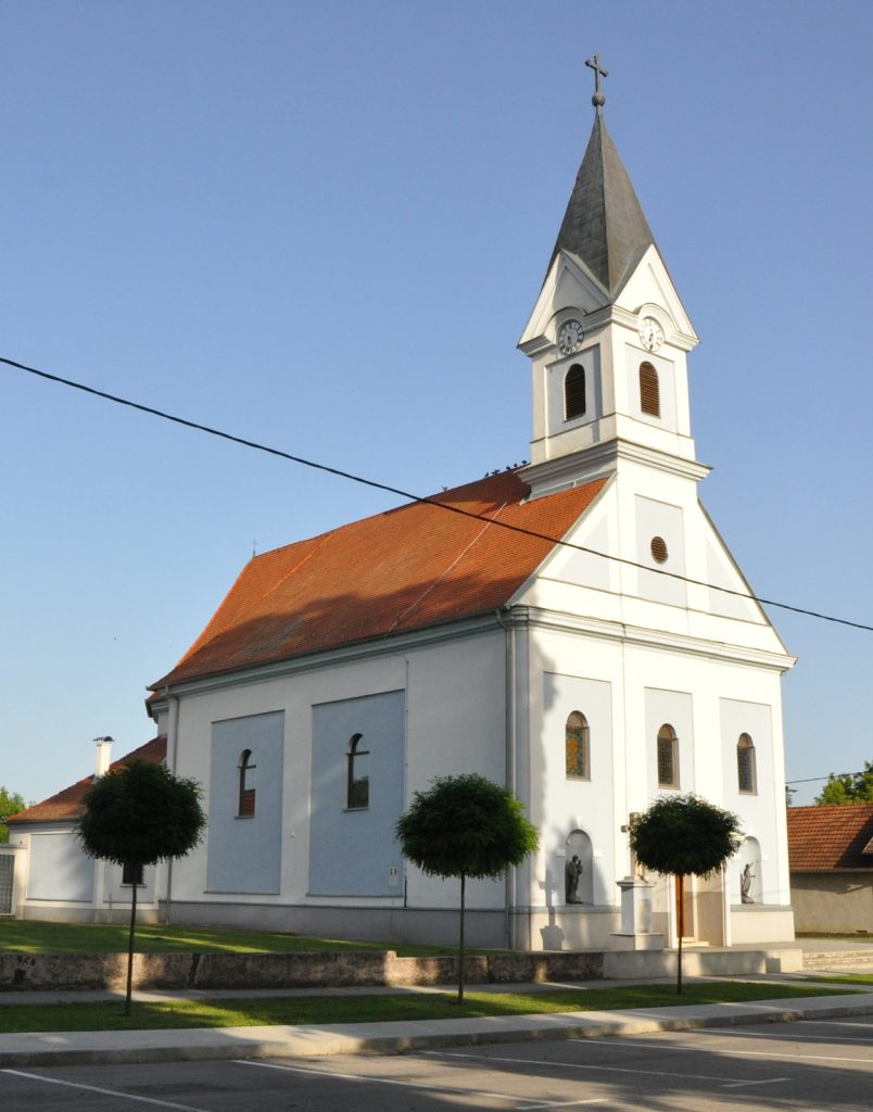 White church with red-tiled roof and tall steeple topped by a cross against a clear blue sky, architectural view from the street.