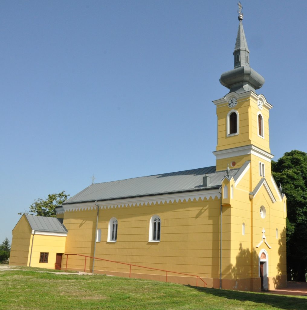 Yellow church with clock tower and tall steeple against a clear blue sky.