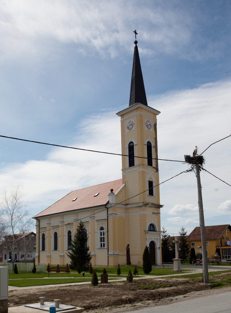 Front view of a yellow church with a tall clock tower and black steeple against a blue sky with clouds