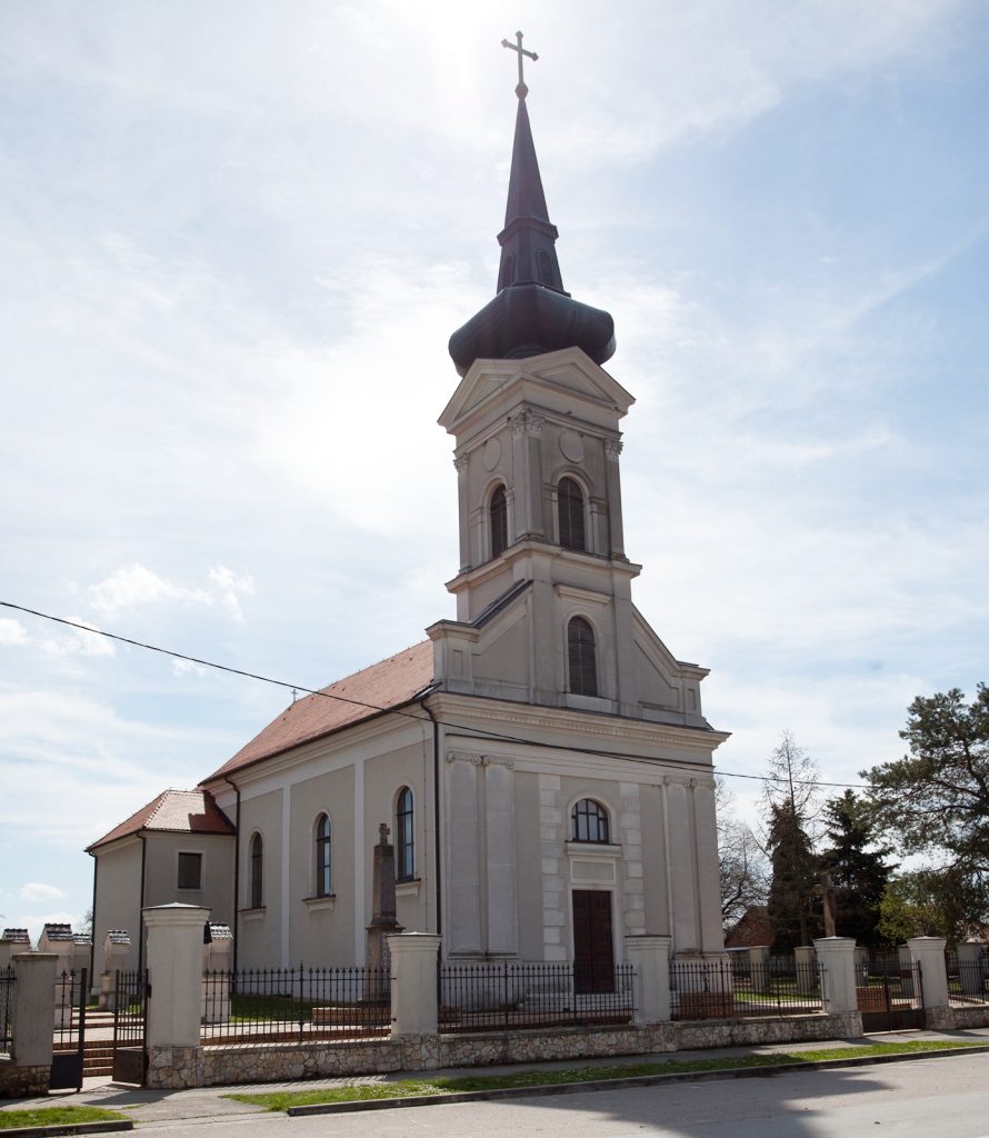 White church with tall dark steeple and cross atop, fenced yard, under a clear blue sky