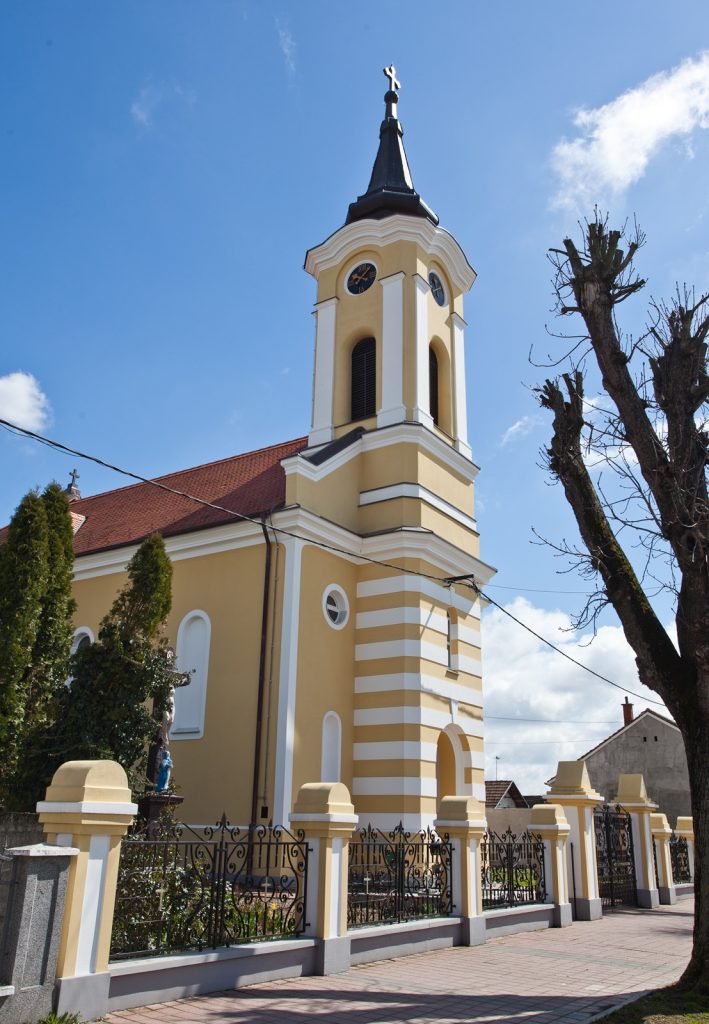 Yellow church with white trim and tall steeple, clock faces on the tower, under a bright blue sky.