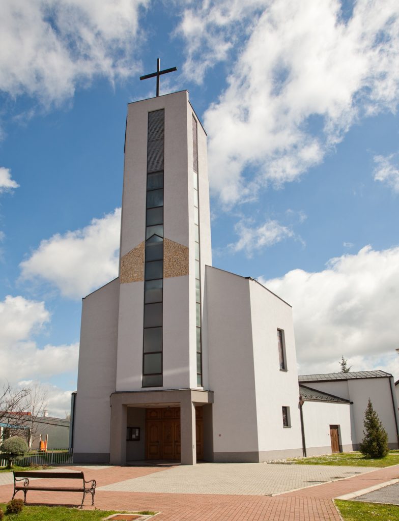 Front view of a modern white church with a tall steeple topped by a cross against a blue sky with clouds