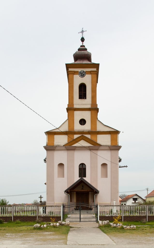 Front view of a tall yellow-and-white church tower with a clock, bell window, and cross atop, fenced by a gate in a rural setting.