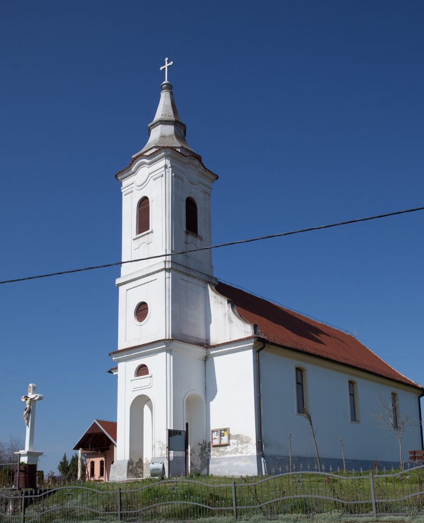 White church with a tall steeple topped by a cross, set against a blue sky, with red-roofed building attached on the right and a metal fence in front.
