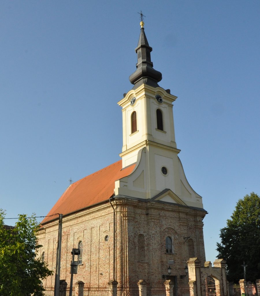 Church with cream tower and dark gray steeple topped by a gold ornament, red-tiled roof, against a clear blue sky, informative shot of a church landmark.