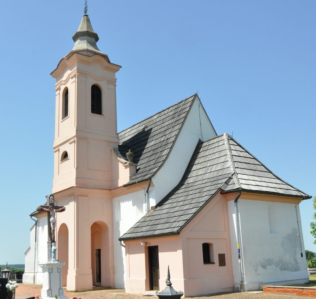 Exterior of a pale pink church with a tall bell tower and arched windows against a clear blue sky; a crucifix statue stands near the entrance.