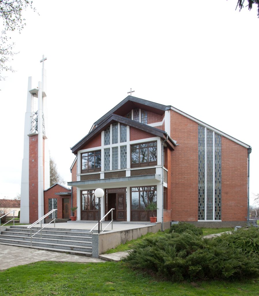 Brick church with large glass-block windows, front stairs, and a cross on the gabled roof, surrounded by greenery.