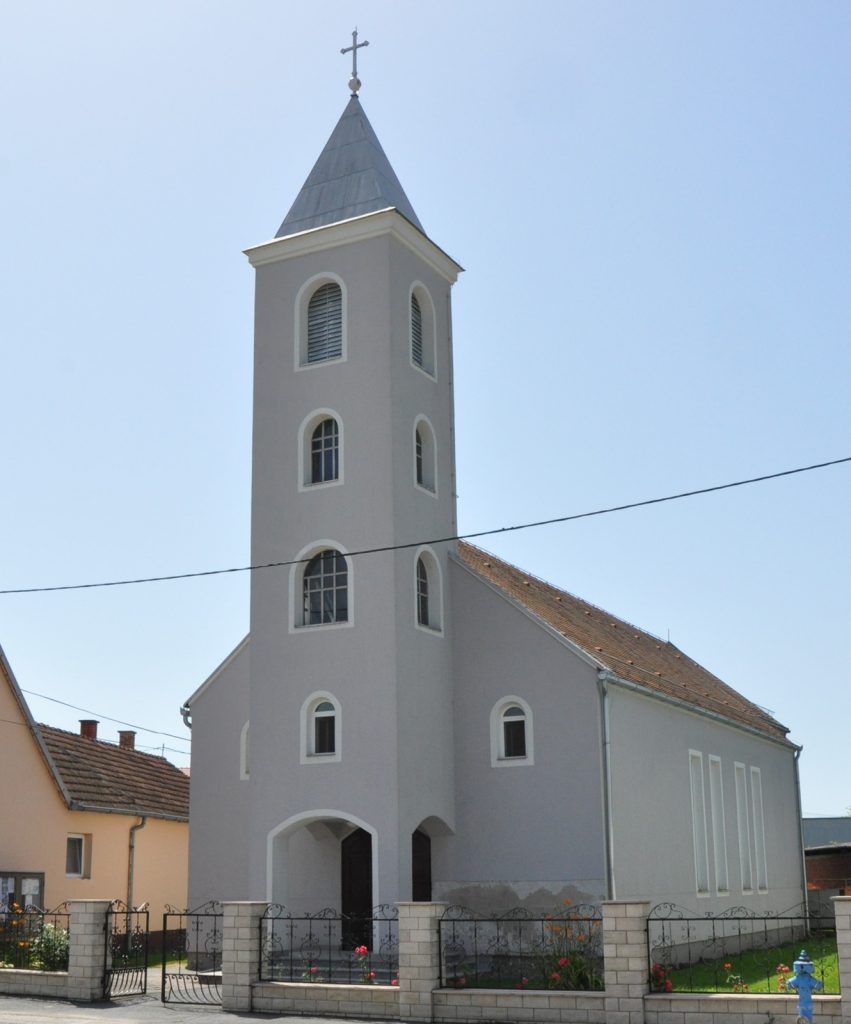 Gray church with a tall square steeple and cross atop, arched white-trimmed windows, and a gated stone fence against a blue sky.