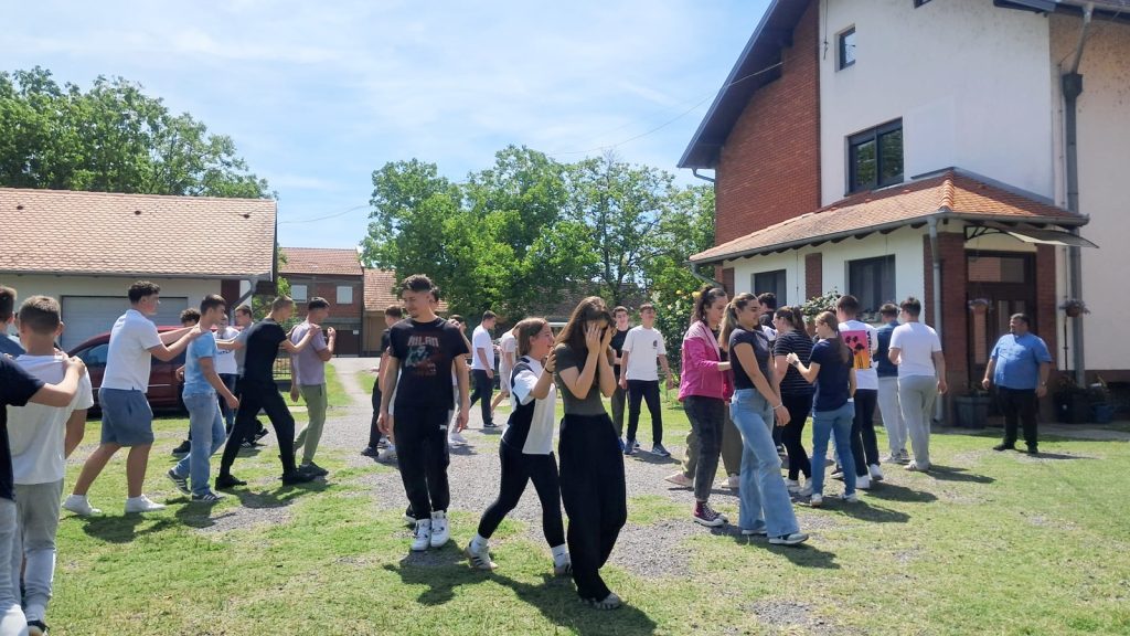 Group of teens outside, paired up and linking arms, participating in an outdoor team activity near a building with a red-tiled roof on a sunny day