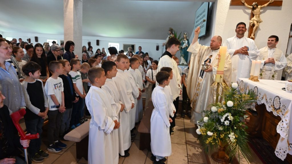 Priests and altar at a church service; children in white robes stand before the altar as a priest raises his hand in blessing.